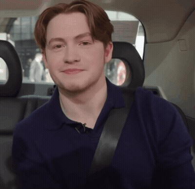 A Young Man Wearing A Blue Shirt And Black Seat Belt Smiles GIF