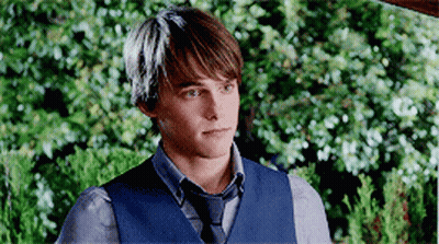 A Young Man Wearing A Blue Vest And Tie Is Standing In Front Of A Bush . GIF