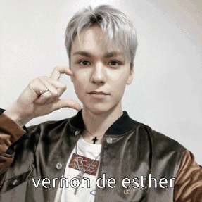 A Young Man Wearing A Brown Jacket Is Making A Heart With His Fingers And The Words Vernon De Esther Below Him GIF