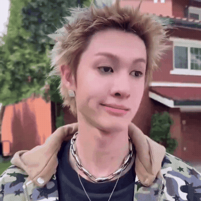 A Young Man Wearing A Camouflage Jacket And A Necklace Looks At The Camera GIF