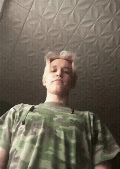 A Young Man Wearing A Camouflage Shirt Is Looking Down At The Camera GIF