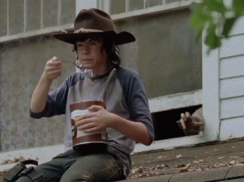 A Young Man Wearing A Cowboy Hat Is Sitting On The Side Of A Building Eating A Bucket Of Food GIF
