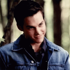 A Young Man Wearing A Denim Jacket And A Black Shirt Is Smiling And Looking At The Camera . GIF