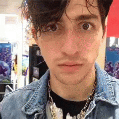 A Young Man Wearing A Denim Jacket And A Necklace Looks At The Camera GIF