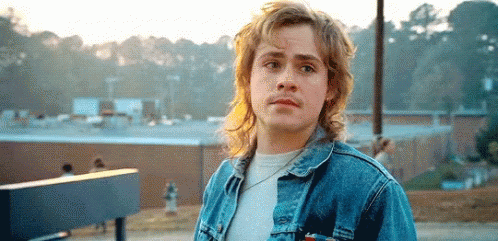 A Young Man Wearing A Denim Jacket And A White Shirt Is Standing In Front Of A Building . GIF