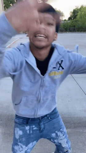 A Young Man Wearing A Gray Hoodie And Blue Jeans Is Standing On A Sidewalk . GIF