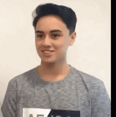 A Young Man Wearing A Gray T-Shirt Is Smiling While Standing In Front Of A White Wall . GIF