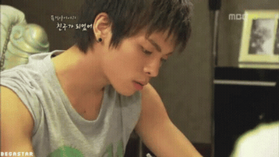 A Young Man Wearing A Gray Tank Top Is Sitting At A Table In Front Of A Mbc Tv GIF
