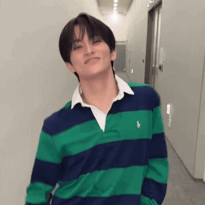 A Young Man Wearing A Green And Blue Striped Shirt Is Standing In A Hallway With His Hands In His Pockets . GIF
