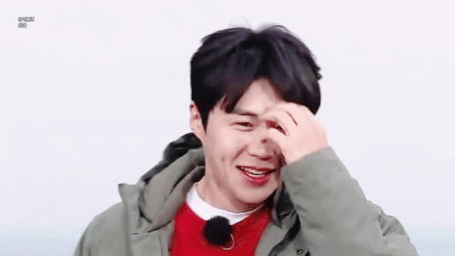 A Young Man Wearing A Green Jacket And A Red Sweater Is Smiling And Covering His Face With His Hand . GIF