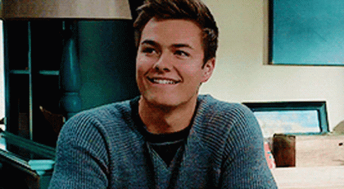 A Young Man Wearing A Grey Sweater Is Smiling While Sitting In Front Of A Lamp . GIF