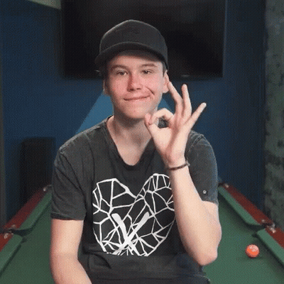 A Young Man Wearing A Hat And A Black Shirt Is Sitting In Front Of A Pool Table Giving An Ok Sign . GIF