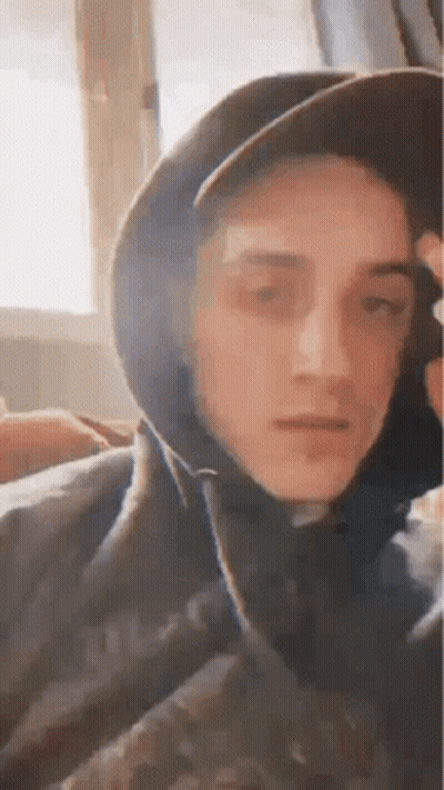 A Young Man Wearing A Hat And A Hoodie Is Sitting In Front Of A Window . GIF