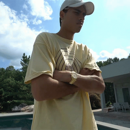 A Young Man Wearing A Hat And A Watch Stands With His Arms Crossed In Front Of A Pool GIF