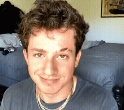A Young Man Wearing A Necklace And A Gray Shirt Smiles For The Camera GIF