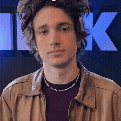 A Young Man Wearing A Necklace And A Leather Jacket Looks At The Camera GIF