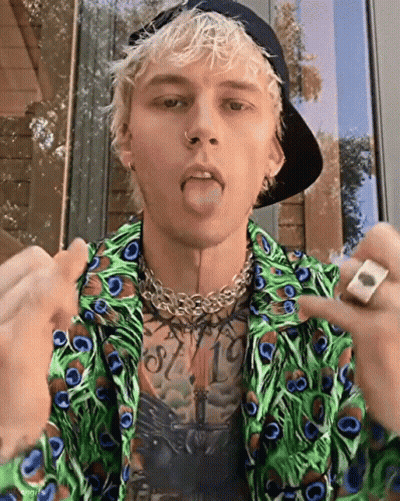 A Young Man Wearing A Peacock Feather Shirt And A Hat Is Sticking His Tongue Out . GIF