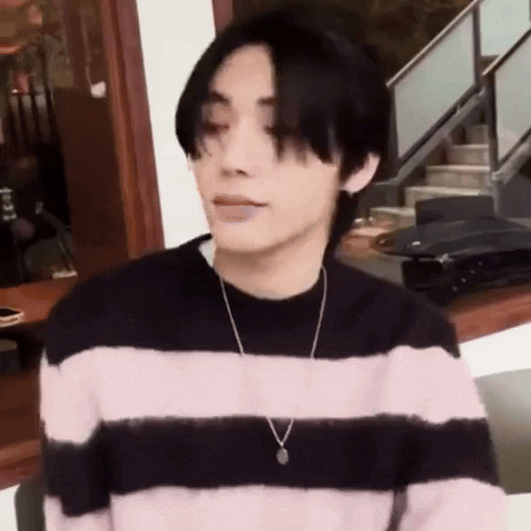 A Young Man Wearing A Pink And Black Striped Sweater And A Necklace Is Sitting In Front Of A Staircase . GIF