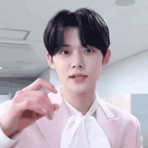 A Young Man Wearing A Pink Jacket And A White Tie Is Making A Heart Shape With His Hand . GIF