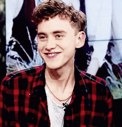A Young Man Wearing A Plaid Shirt And A Necklace Is Smiling GIF