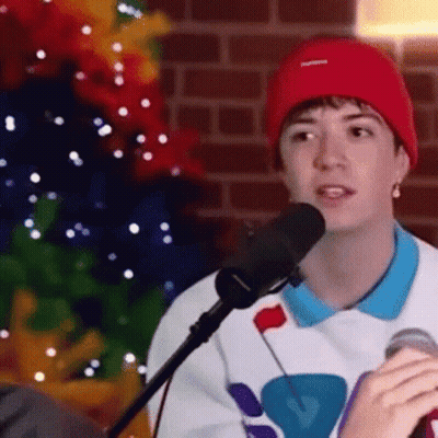 A Young Man Wearing A Red Beanie Is Singing Into A Microphone While Sitting In Front Of A Christmas Tree . GIF