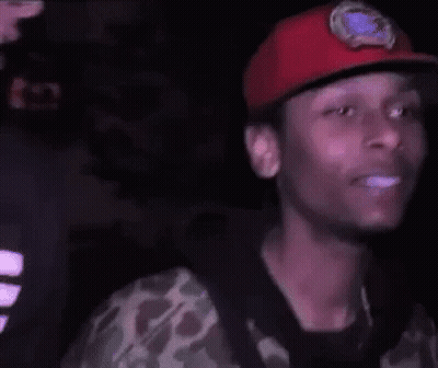 A Young Man Wearing A Red Hat And A Camouflage Shirt Is Smiling In The Dark . GIF