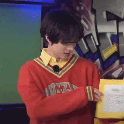 A Young Man Wearing A Red Sweater And A Yellow Shirt Is Holding A Piece Of Paper . GIF