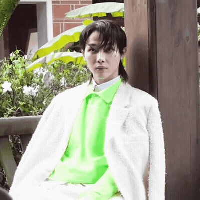 A Young Man Wearing A White Jacket And A Neon Green Sweater Is Sitting On A Bench . GIF