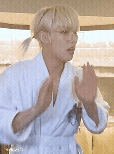 A Young Man Wearing A White Robe And Glasses Is Standing In A Room . GIF