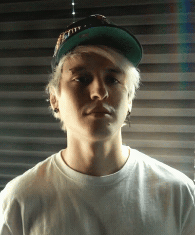 A Young Man Wearing A White Shirt And A Green Hat Looks At The Camera With A Serious Look On His Face GIF