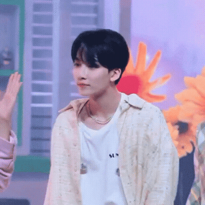 A Young Man Wearing A White Shirt And A Pearl Necklace Is Standing In Front Of A Bunch Of Flowers . GIF