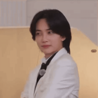 A Young Man Wearing A White Suit And Black Tie Is Sitting In Front Of A Piano . GIF