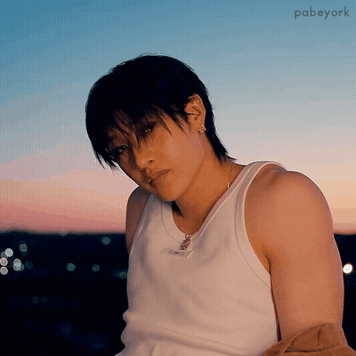 A Young Man Wearing A White Tank Top And Earrings Looks At The Camera . GIF