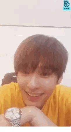A Young Man Wearing A Yellow Shirt Is Smiling While Holding A Watch In Front Of A Vlive Logo Sticker