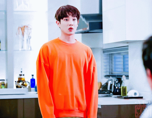 A Young Man Wearing An Orange Sweater Stands In A Kitchen GIF