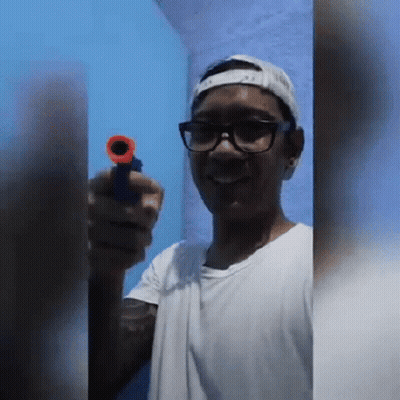 A Young Man Wearing Glasses And A Baseball Cap Is Holding A Toy Gun . GIF