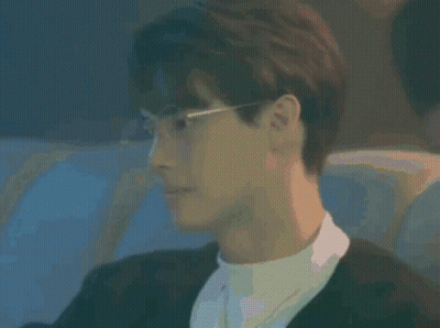 A Young Man Wearing Glasses And A Black Jacket Is Sitting In A Dark Room . GIF