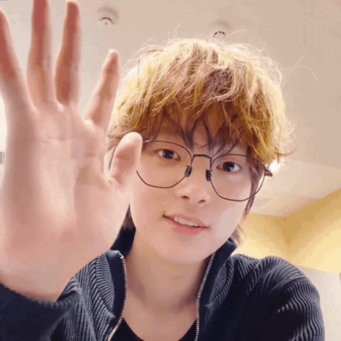 A Young Man Wearing Glasses And A Black Sweater Is Waving His Hand GIF