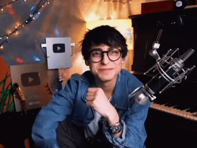 A Young Man Wearing Glasses And A Denim Jacket Is Sitting In Front Of A Microphone And A Piano . GIF