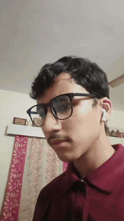 A Young Man Wearing Glasses And A Maroon Shirt Is Looking At The Camera GIF