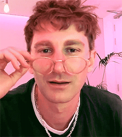 A Young Man Wearing Glasses And A Necklace Is Looking At The Camera . GIF
