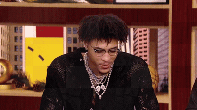 A Young Man Wearing Glasses And A Necklace Is Sitting In Front Of A Display Case . GIF