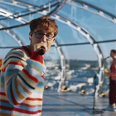 A Young Man Wearing Glasses And A Striped Sweater Sings Into A Microphone GIF