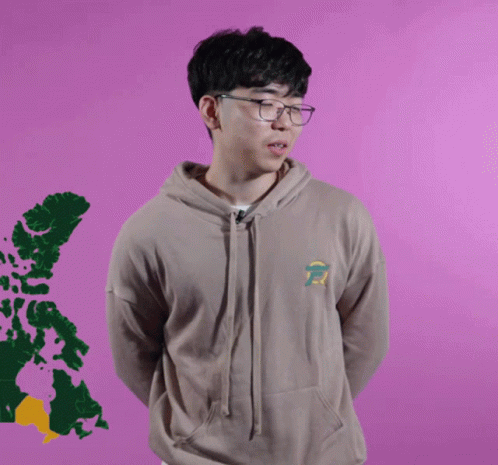 A Young Man Wearing Glasses And A Tan Hoodie Stands In Front Of A Purple Background GIF
