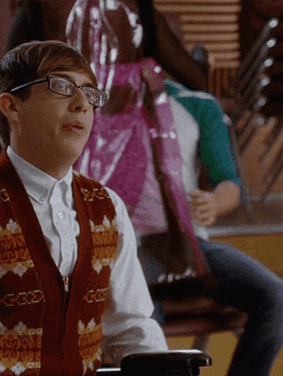 A Young Man Wearing Glasses And A Vest That Says ' A ' On It Sits In A Chair GIF