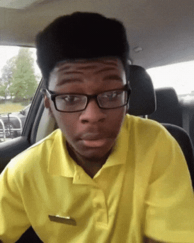 A Young Man Wearing Glasses And A Yellow Shirt Is Sitting In The Driver 'S Seat Of A Car GIF
