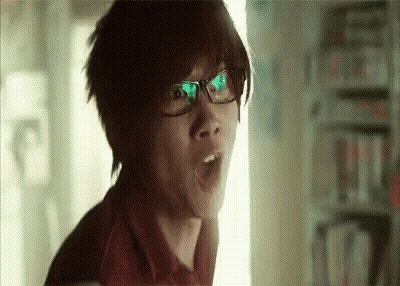 A Young Man Wearing Glasses Is Making A Surprised Face In A Library . GIF