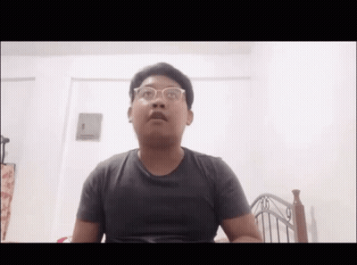 A Young Man Wearing Glasses Is Sitting In Front Of A Bed . GIF