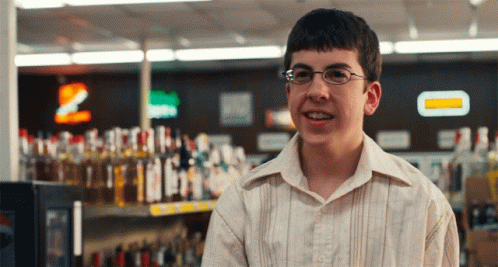 A Young Man Wearing Glasses Is Smiling In Front Of A Shelf Of Liquor GIF