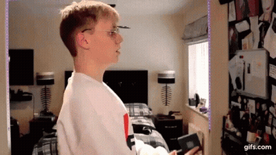 A Young Man Wearing Glasses Is Standing In A Bedroom Next To A Bed And A Window . GIF
